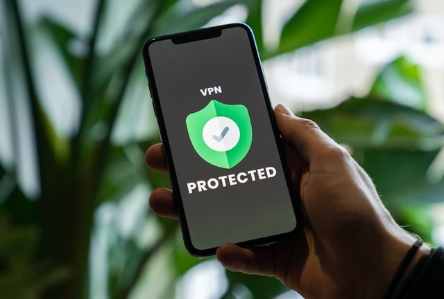 Uma mão segura um telefone celular, e a tela mostra um crachá com as palavras "VPN PROTECTED".