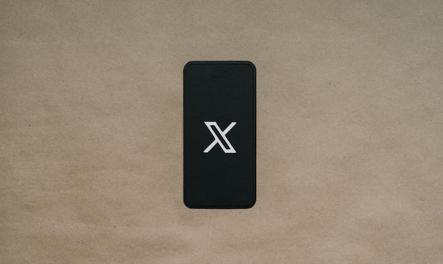 A picture of a white X logo on the screen of a black smartphone placed on brown paper.